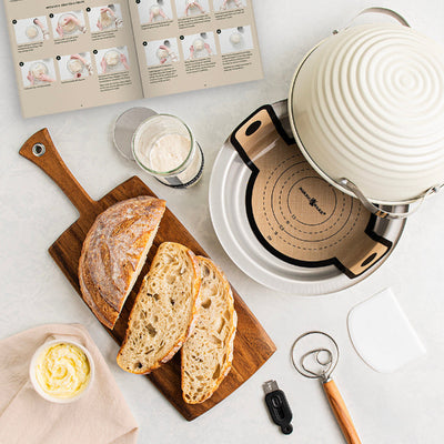 Loaf of bread on a wooden cutting board with a pot and kitchen items on a light surface.