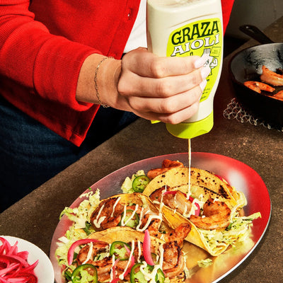 A person in a red cardigan squeezes a white sauce from a bottle labeled GRAZA AIOLI onto a plate of grilled tacos on a red oval platter.