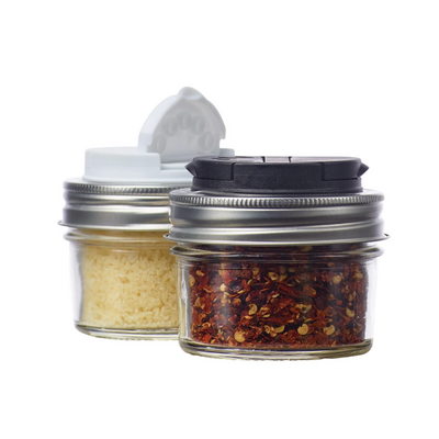 Two small clear glass spice jars with metal screw-top lids and flip-top dispensers, one filled with white seasoning and one with red pepper flakes, on a white background.
