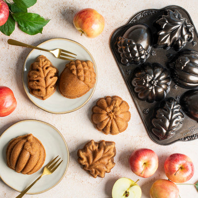 Acorn-shaped and leaf-shaped pumpkin breads displayed on plates with gold forks, alongside a black metal loaf mold and scattered red and yellow apples on a light stone surface.