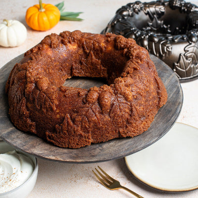 A ring-shaped brown bundt cake sits on a dark round serving platter, with a decorative metal bundt mold, small pumpkins, plates, and a fork arranged around it on a light surface.