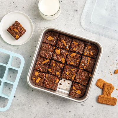 Brownies in a baking tray with a slice on a plate, glass of milk, and Biscoff cookies on a gray surface.