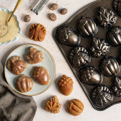 Acorn-shaped metal mold tray filled with baked acorn cookies, alongside plated acorn cookies and scattered acorns on a light stone surface.
