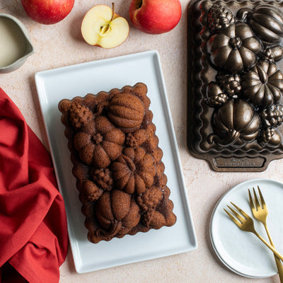 A rectangular loaf cake with a decorative pumpkin and acorn mold pattern sits on a white plate, styled with whole and sliced apples, a red cloth, gold forks, and the dark metal mold on a light beige surface.