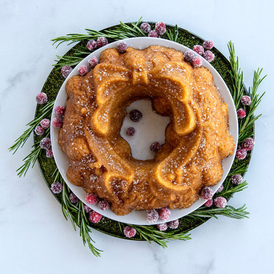 A golden-brown bundt cake with a white glaze, dusted with powdered sugar, and garnished with cranberries, set on a green plate with rosemary sprigs on a white marble surface.