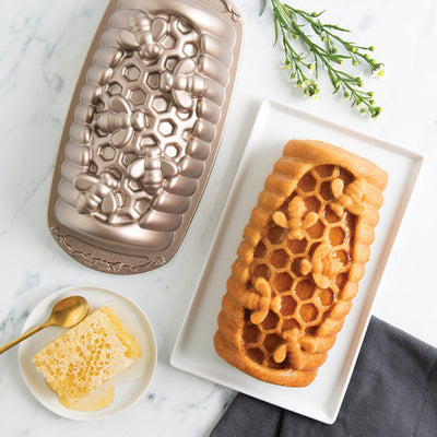 Honeycomb-shaped baked good next to a honeycomb mold and plate of honey on a marble surface.