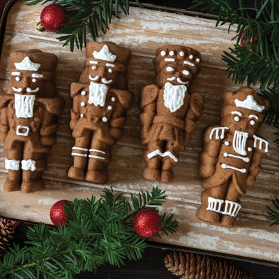 Four decorated gingerbread nutcracker cookies arranged on a rustic wooden board with evergreen branches and red ornaments around them.