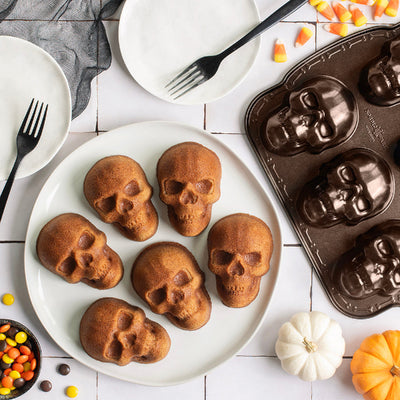 Six skull-shaped mini loaves of bread arranged on a white plate, styled with a chocolate skull mold, candy corn, small pumpkins, and black forks on a white tile surface.
