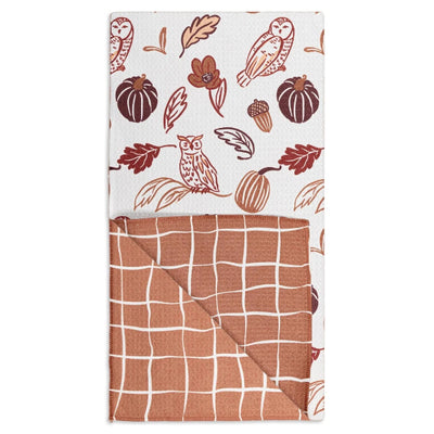 A folded waffle-weave tea towel with a warm autumn pattern. The top half shows a white background with illustrations of owls, pumpkins, acorns, and leaves in terracotta, brown, and rust.